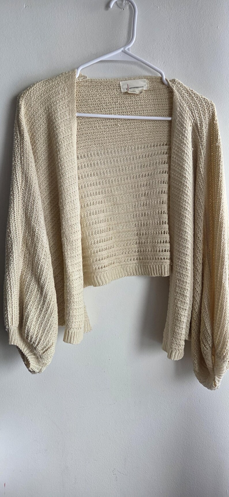 May include: Cream-colored open-front cardigan with long, voluminous sleeves. The knitwear features a textured pattern and is displayed on a white hanger against a white wall. A tag is visible on the back of the neck.