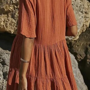 May include: Rust-colored tiered dress with short sleeves. The dress has a textured, crinkled fabric and a relaxed fit. The model is wearing a gold bracelet. The background is a rocky, neutral-toned environment.