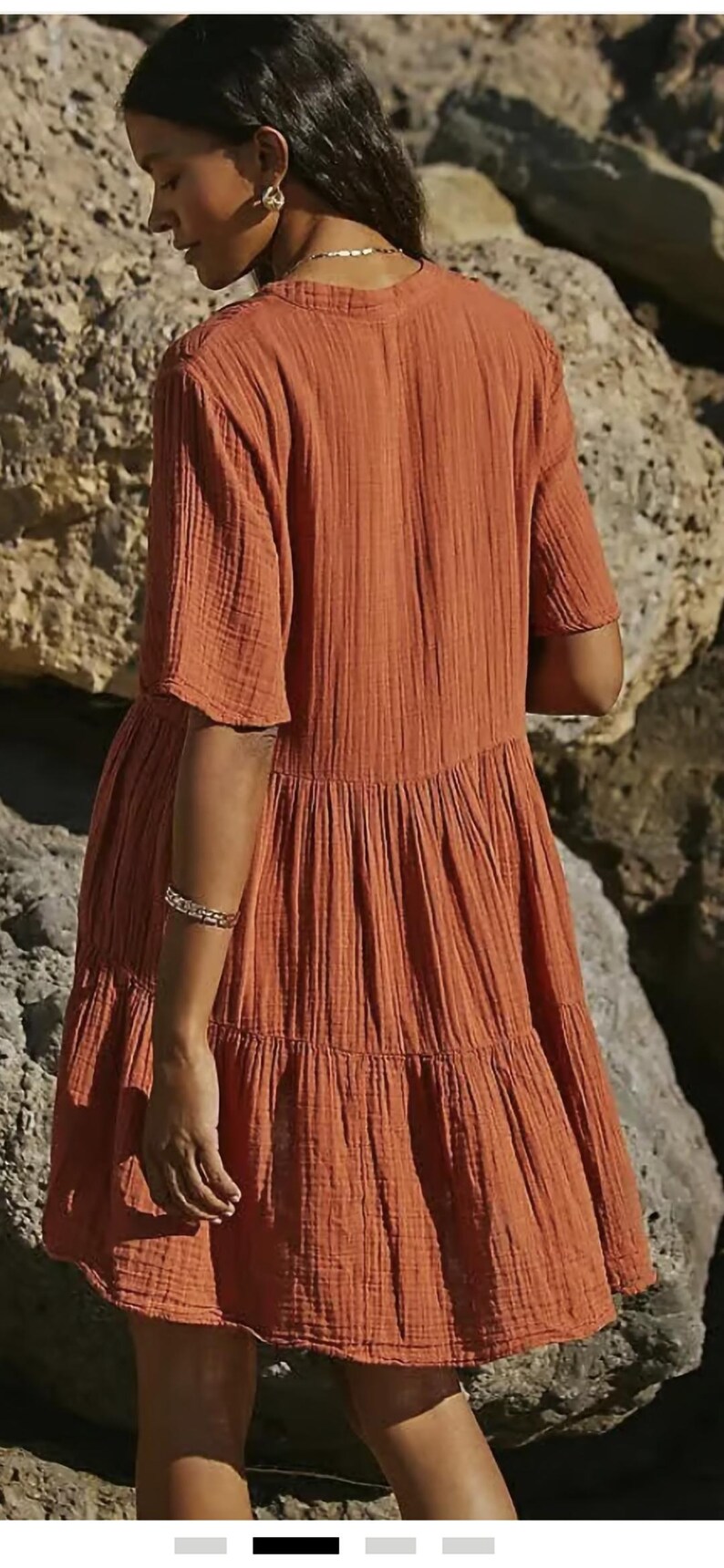 May include: Rust-colored tiered dress with short sleeves. The dress has a textured, crinkled fabric and a relaxed fit. The model is wearing a gold bracelet. The background is a rocky, neutral-toned environment.
