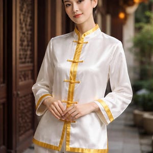May include: A white silk top and pants set with gold trim. The long-sleeved top features a mandarin collar and gold frog closures down the front. Gold trim accents the collar, cuffs, and front edges. The outfit is a traditional style.