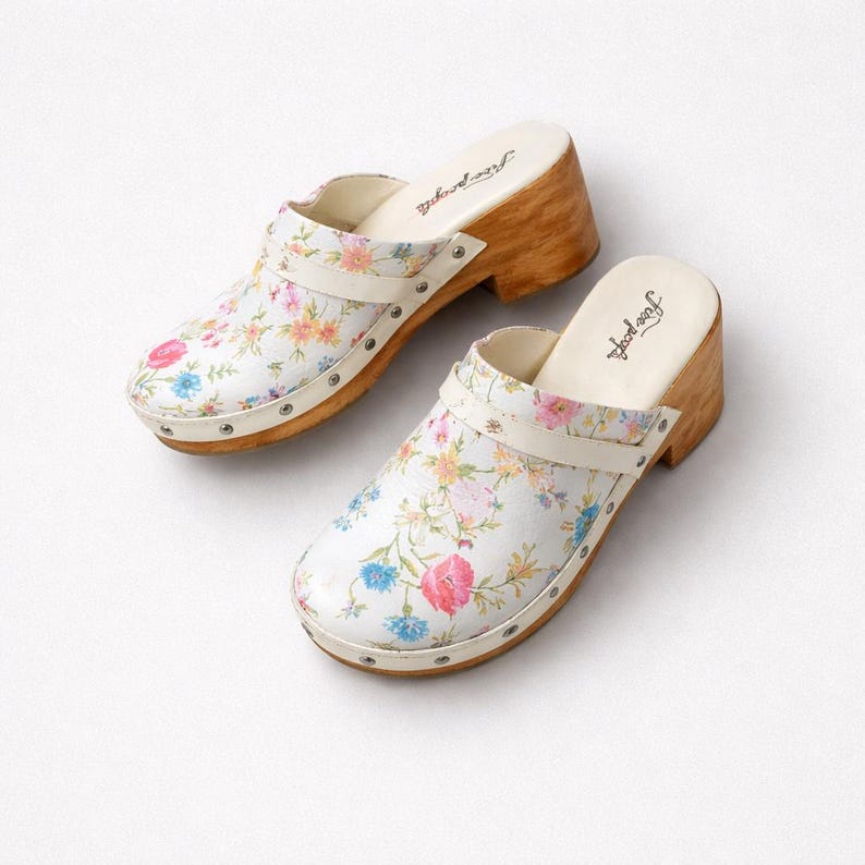 May include: A pair of white clogs with a floral pattern featuring pink, blue, and yellow flowers. The clogs have wooden heels, cream-colored straps, and are adorned with small silver studs. The brand name "Free People" is printed on the insole.
