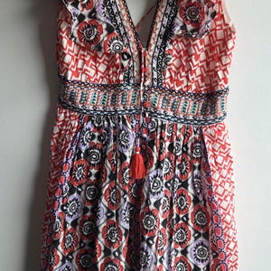 May include: A sleeveless romper with a V-neck and a floral pattern in red, black, and purple. The romper features a tassel detail and ruffled sleeves. The waist is cinched with an embroidered band.