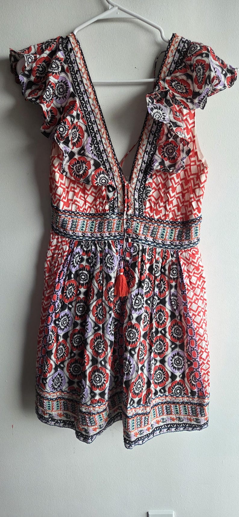May include: A sleeveless romper with a V-neck and a floral pattern in red, black, and purple. The romper features a tassel detail and ruffled sleeves. The waist is cinched with an embroidered band.