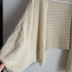 May include: Cream-colored open-front cardigan with wide sleeves. The knitwear features a textured pattern on the sleeves and a more open, airy weave on the body. The garment is hanging on a white hanger against a plain wall.