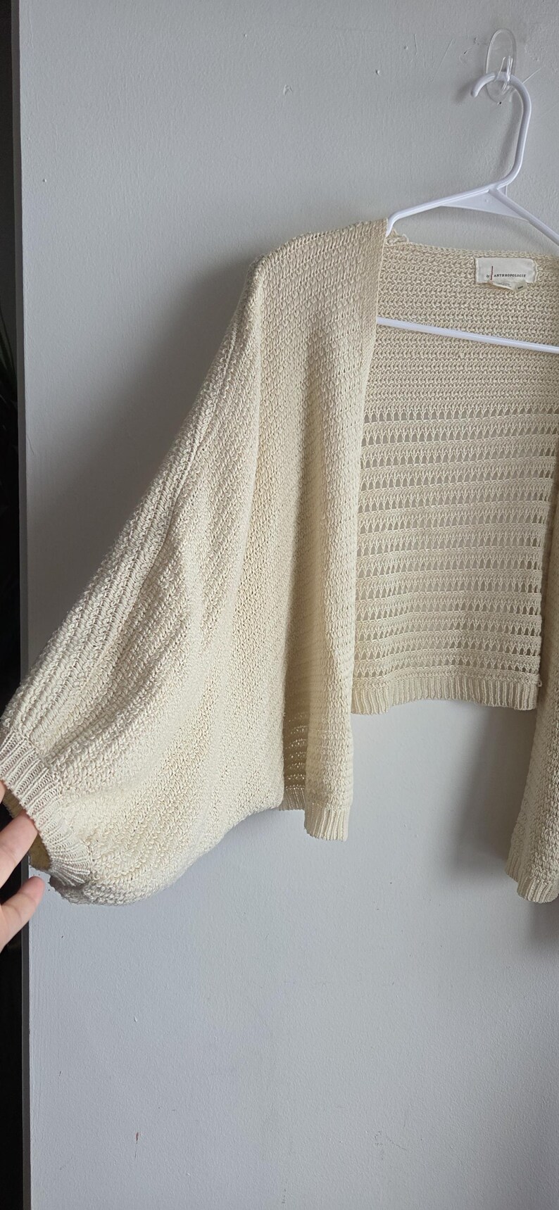 May include: Cream-colored open-front cardigan with wide sleeves. The knitwear features a textured pattern on the sleeves and a more open, airy weave on the body. The garment is hanging on a white hanger against a plain wall.