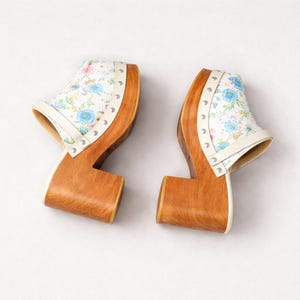 May include: A pair of wooden clogs with a white floral patterned upper. The clogs have a light brown wooden base and a white leather trim. The upper features a blue, pink, and green floral design. Silver studs secure the upper to the wooden base.