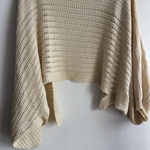 May include: Cream-colored, open-weave knit shrug with wide sleeves, hanging on a white hanger. The shrug has a cropped length and a loose, flowing design. The knit pattern features a combination of solid and open sections.