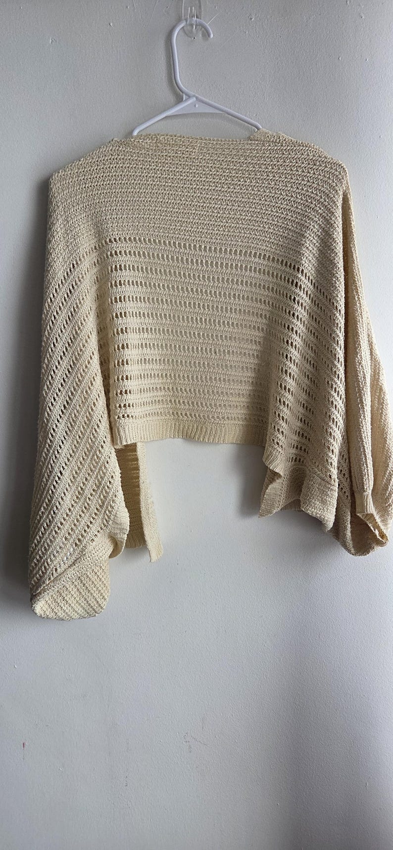 May include: Cream-colored, open-weave knit shrug with wide sleeves, hanging on a white hanger. The shrug has a cropped length and a loose, flowing design. The knit pattern features a combination of solid and open sections.