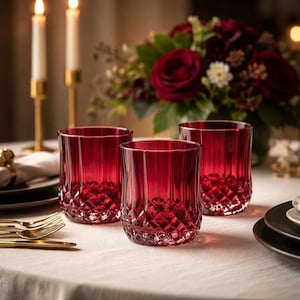 May include: Three ruby red drinking glasses with a diamond-cut base and vertical lines. The glasses are on a white tablecloth, with a floral arrangement and lit candles in the background. The glasses are empty.