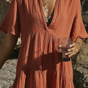 May include: Rust-colored, short-sleeved dress with a V-neck. The dress has a tiered skirt and is made of a textured fabric. A person is wearing the dress and holding a glass of water. A silver necklace is visible.