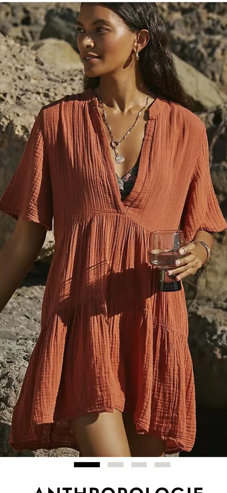 May include: Rust-colored, short-sleeved dress with a V-neck. The dress has a tiered skirt and is made of a textured fabric. A person is wearing the dress and holding a glass of water. A silver necklace is visible.