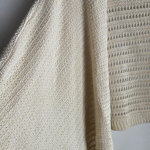 May include: Cream-colored open-front cardigan sweater. The knitwear features a textured pattern on the sleeves and a more open, lacy design on the front panels. The sweater is hanging on a white hanger against a plain wall.