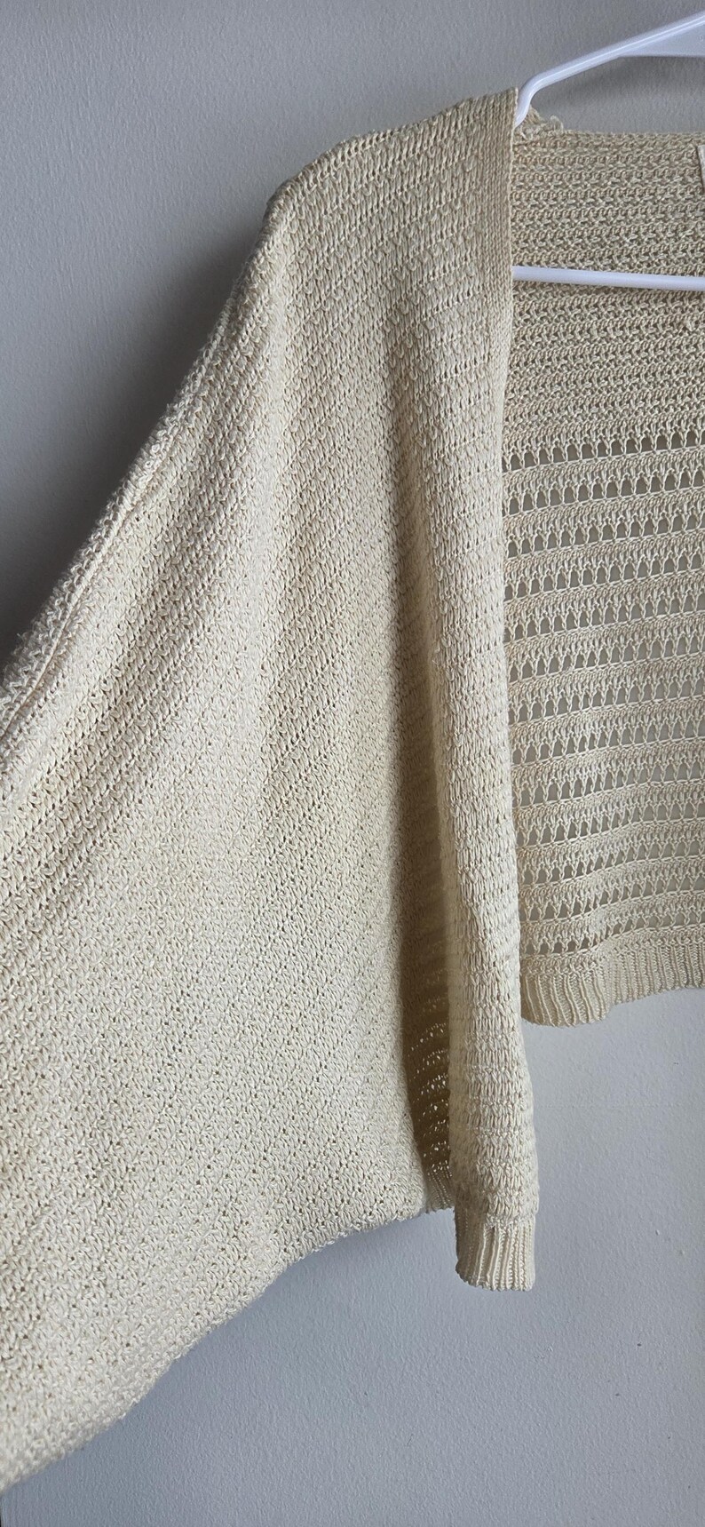 May include: Cream-colored open-front cardigan sweater. The knitwear features a textured pattern on the sleeves and a more open, lacy design on the front panels. The sweater is hanging on a white hanger against a plain wall.