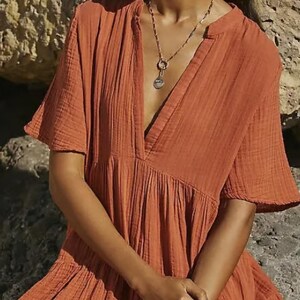 May include: Rust-colored short-sleeve dress with a V-neckline. The dress has a tiered skirt and a relaxed fit. A gold-toned necklace and bracelet are visible. The fabric appears to be a lightweight, textured material.