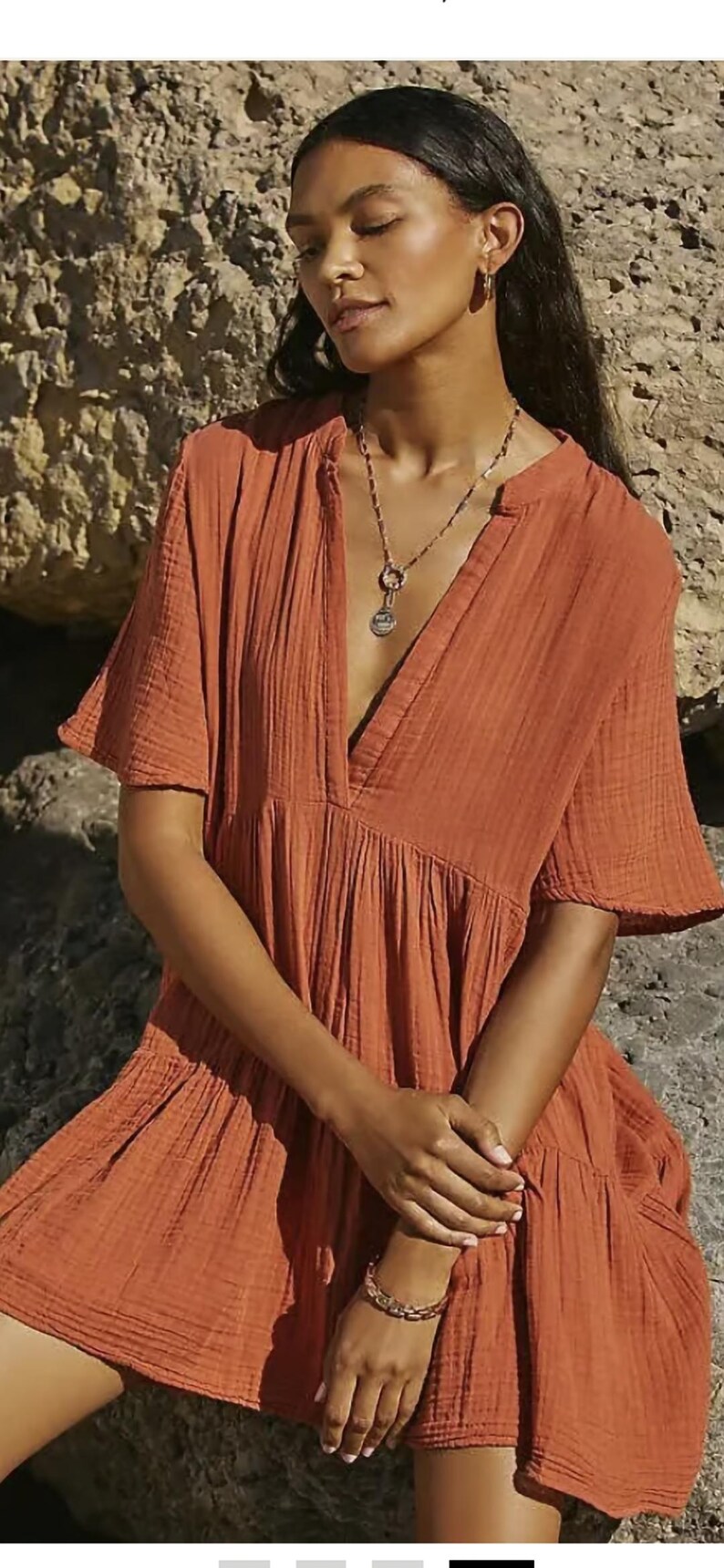 May include: Rust-colored short-sleeve dress with a V-neckline. The dress has a tiered skirt and a relaxed fit. A gold-toned necklace and bracelet are visible. The fabric appears to be a lightweight, textured material.