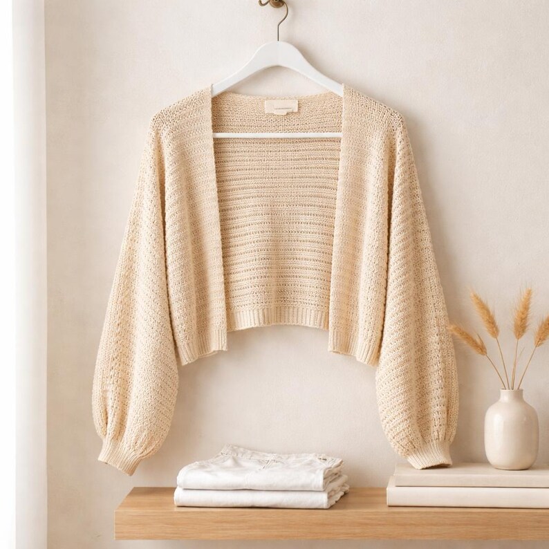 May include: A cream-colored, open-front bolero sweater with long, voluminous sleeves. The sweater is made of a textured knit fabric and hangs on a white hanger. A wooden shelf below holds folded white linens and a vase with dried plants.