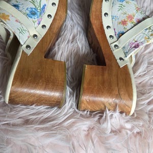 May include: Pair of wooden clogs with floral print uppers. The clogs have a light wood base and a white strap with silver studs. The floral pattern includes blue, pink, and yellow flowers on a white background.