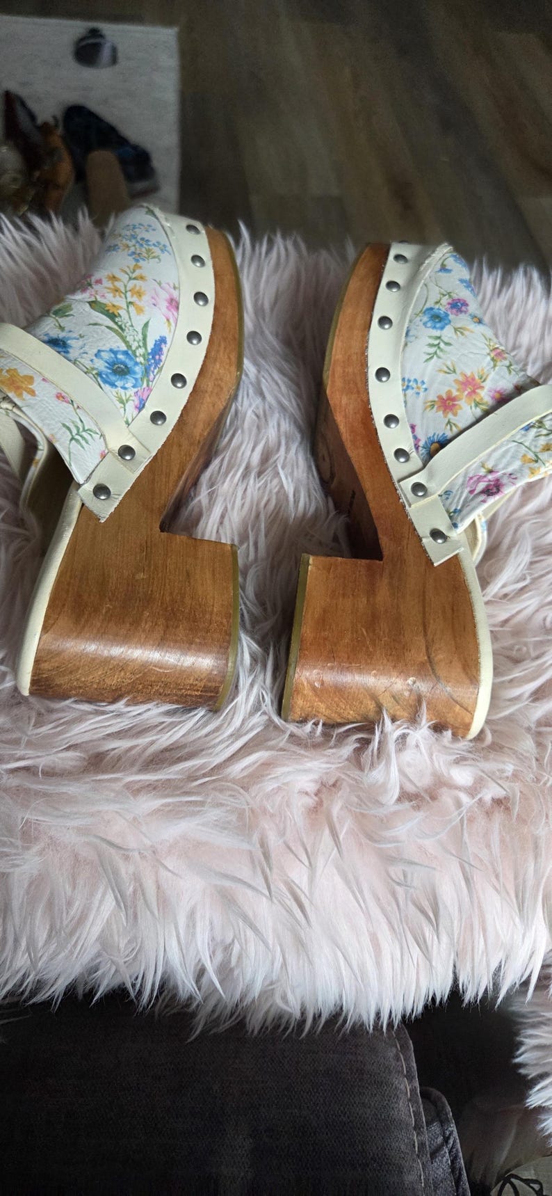 May include: Pair of wooden clogs with floral print uppers. The clogs have a light wood base and a white strap with silver studs. The floral pattern includes blue, pink, and yellow flowers on a white background.