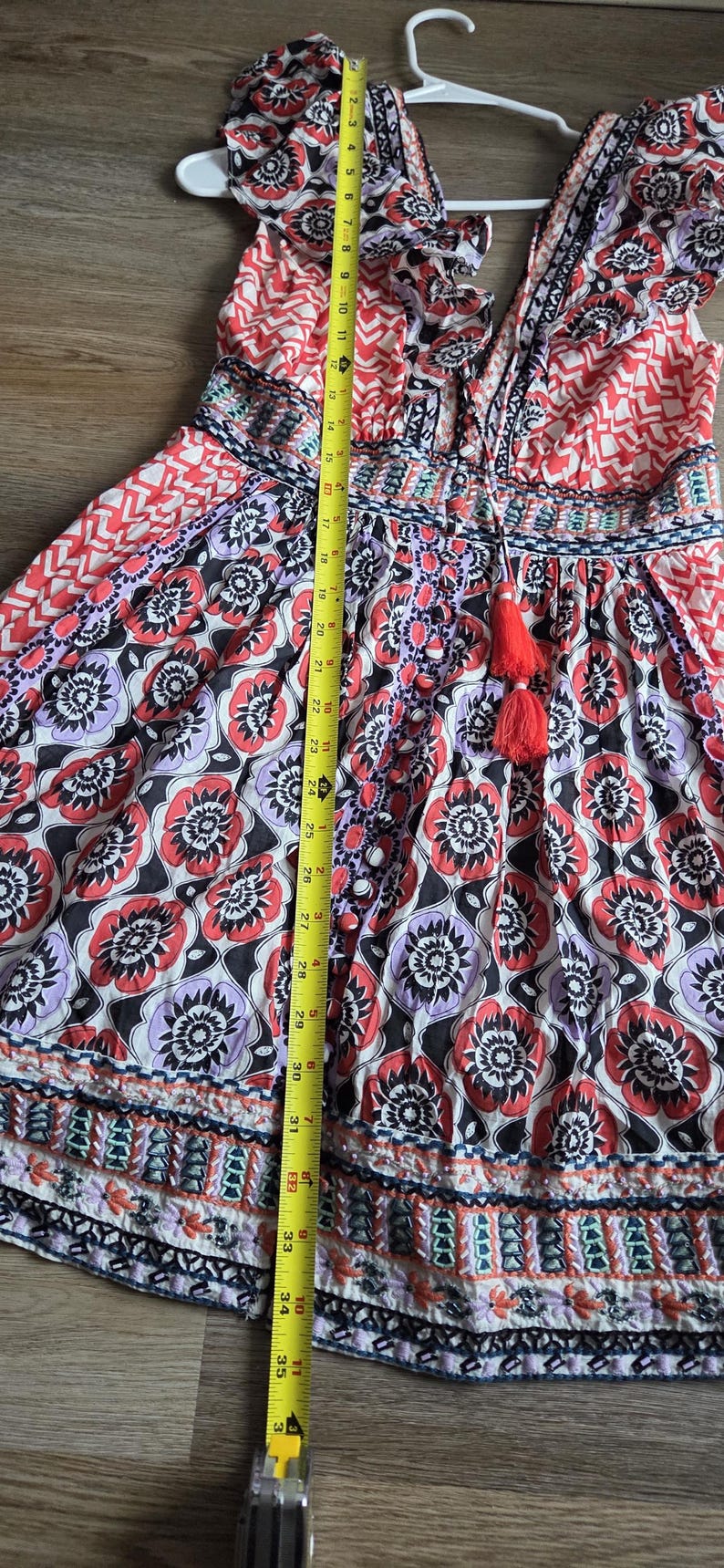 May include: A patterned dress with a V-neck and tassel detail. The dress features a floral print in red, black, and purple, and measures approximately 89 cm in length. It is hanging on a white hanger.