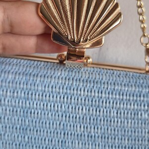 Puede incluir: Un bolso de mano tejido en color azul claro con un marco de metal dorado y un cierre prominente en forma de concha marina dorada. El bolso incluye una fina correa de cadena de metal dorado unida al marco superior, con un aspecto estructurado.