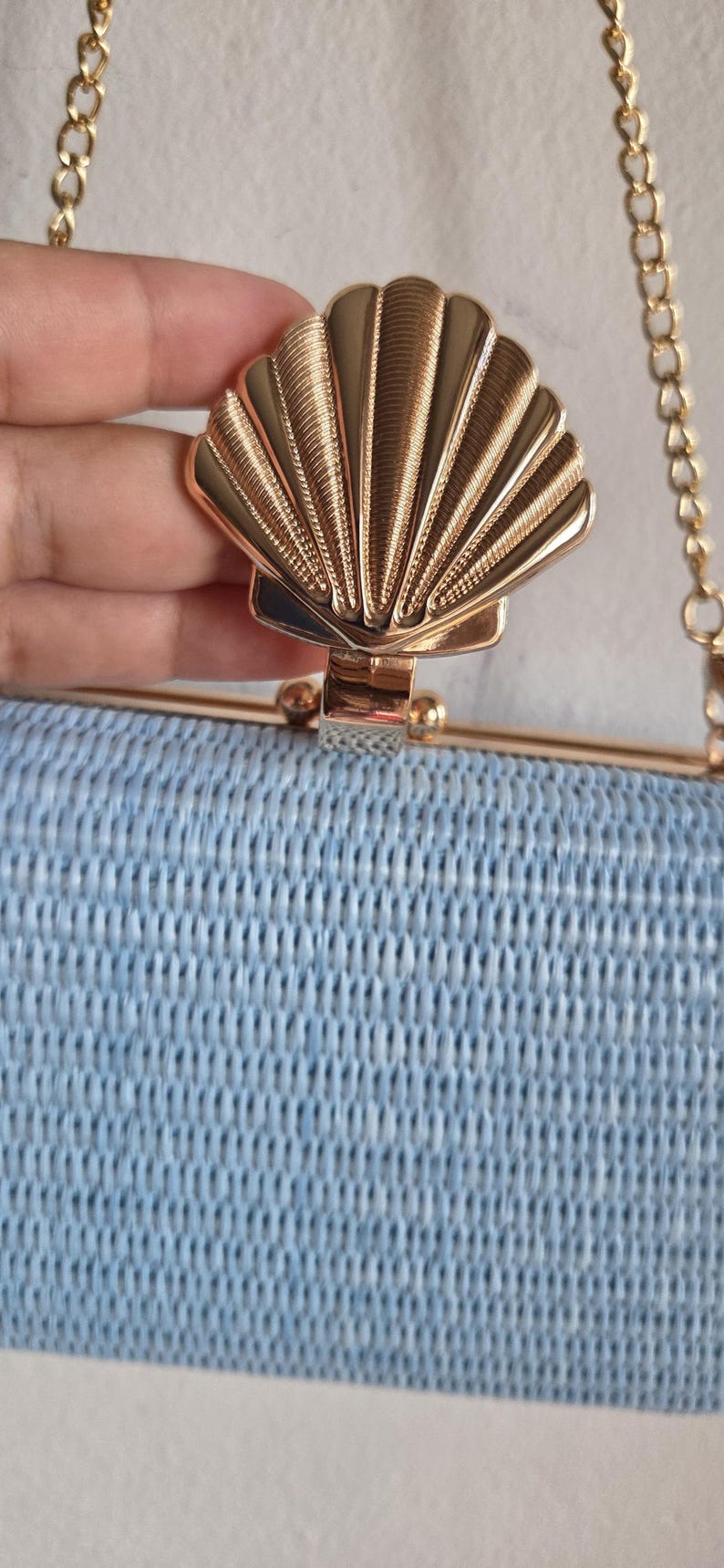 Puede incluir: Un bolso de mano tejido en color azul claro con un marco de metal dorado y un cierre prominente en forma de concha marina dorada. El bolso incluye una fina correa de cadena de metal dorado unida al marco superior, con un aspecto estructurado.