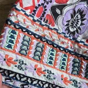 May include: Close-up of a section of fabric with detailed embroidery and beadwork. The design includes floral and geometric patterns in red, white, purple, black, and orange. The fabric is likely part of a garment.
