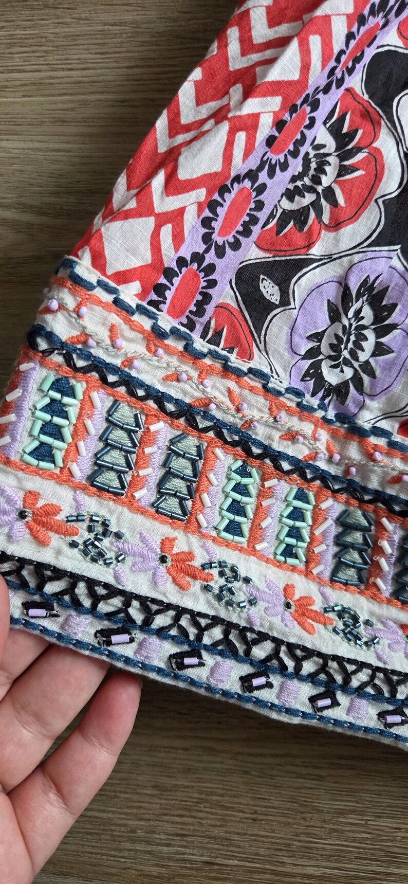 May include: Close-up of a section of fabric with detailed embroidery and beadwork. The design includes floral and geometric patterns in red, white, purple, black, and orange. The fabric is likely part of a garment.