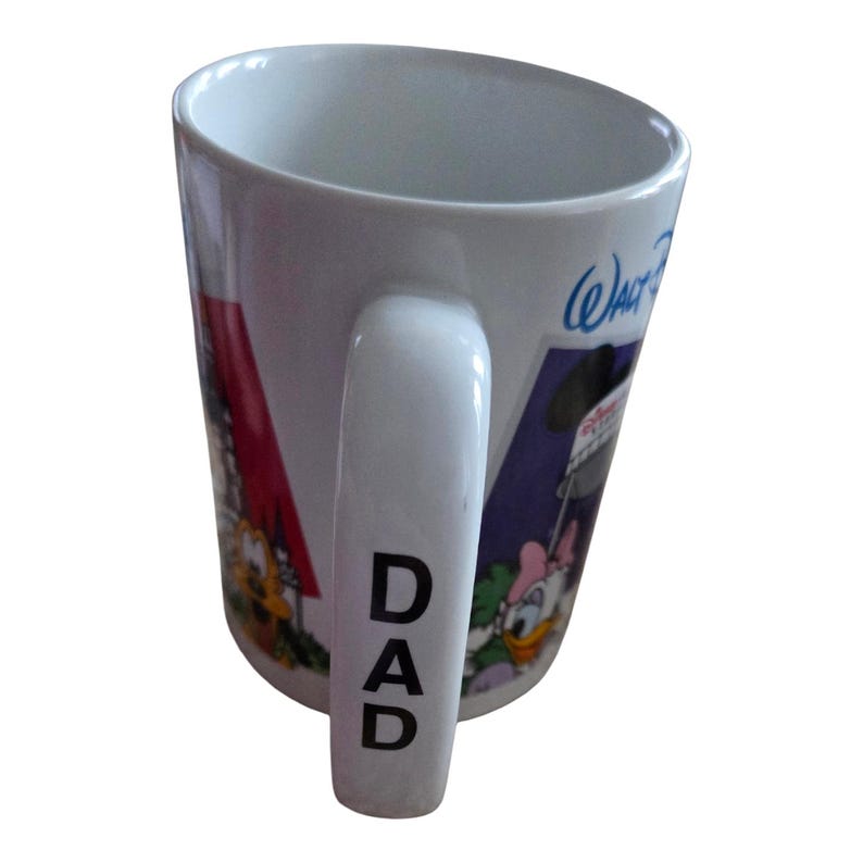May include: White ceramic mug with a curved handle, featuring cartoon characters and the word "DAD" printed vertically on the handle. The mug has a colorful design with the words "Walt Disney" in blue.