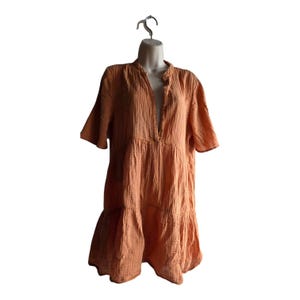 May include: Peach-colored short-sleeve dress with a tiered skirt and a V-neckline. The dress is made of a textured fabric and hangs on a white mannequin. The dress has a relaxed fit and a casual style.