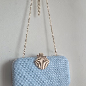 Puede incluir: Un bolso de mano rectangular de color azul claro con una superficie tejida texturizada. El bolso cuenta con un cierre de metal dorado en forma de concha en el centro y una fina correa de cadena de metal dorado. El dise&ntilde;o es estructurado.