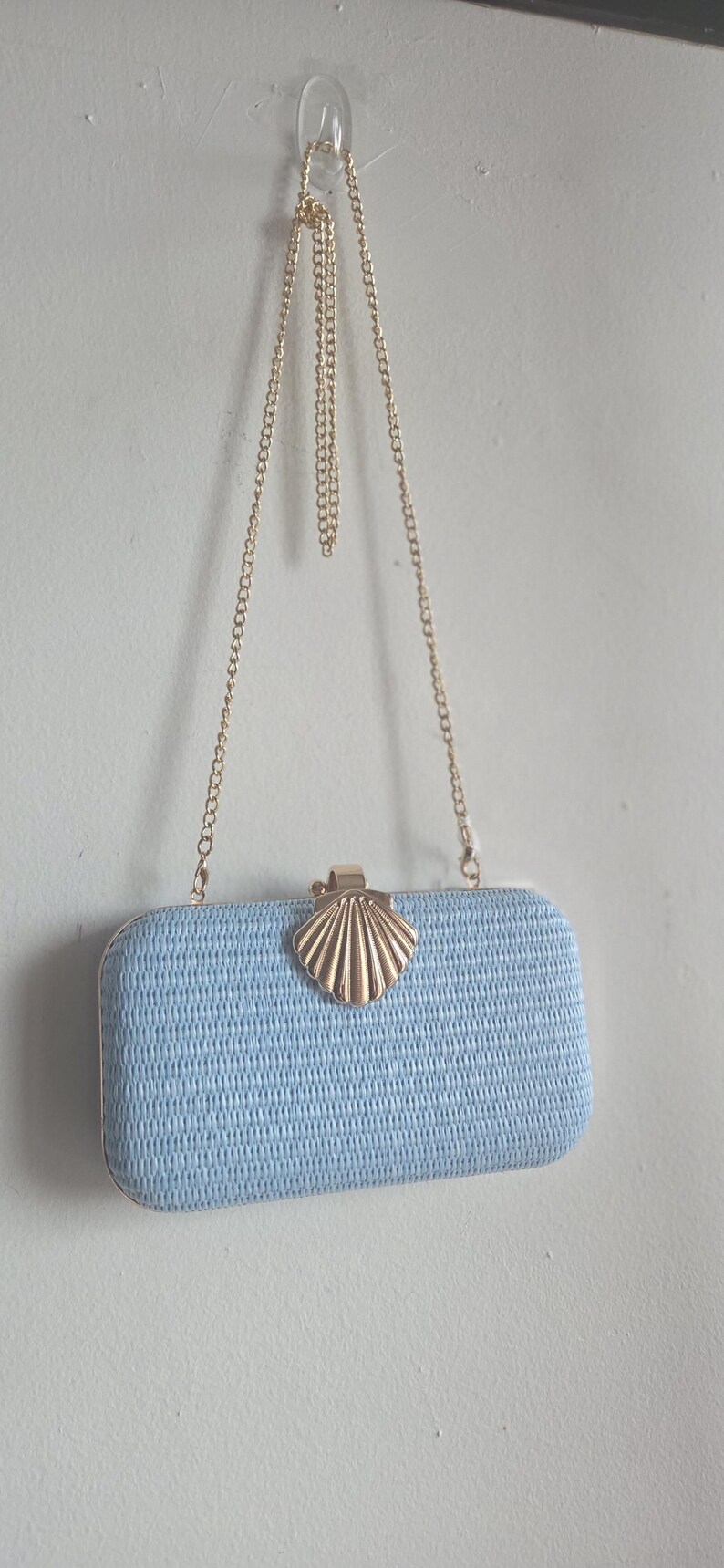 Puede incluir: Un bolso de mano rectangular de color azul claro con una superficie tejida texturizada. El bolso cuenta con un cierre de metal dorado en forma de concha en el centro y una fina correa de cadena de metal dorado. El dise&ntilde;o es estructurado.
