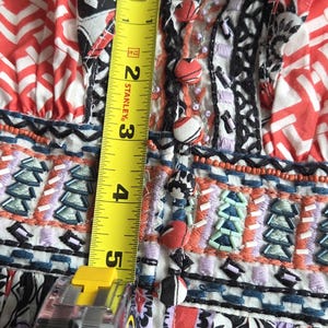 May include: Close-up of a patterned fabric with a tape measure showing a measurement of approximately 13 cm. The fabric features red, white, black, and purple floral and geometric designs, with embroidered details and decorative buttons.