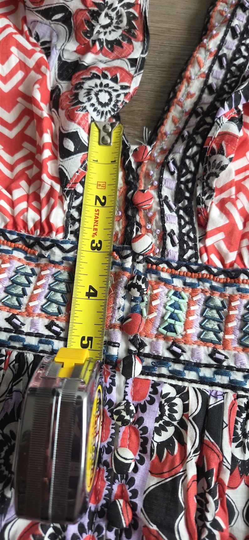 May include: Close-up of a patterned fabric with a tape measure showing a measurement of approximately 13 cm. The fabric features red, white, black, and purple floral and geometric designs, with embroidered details and decorative buttons.