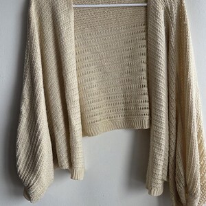 May include: A cream-colored, open-front cardigan with long, voluminous sleeves. The knitwear features a textured pattern and is displayed on a white hanger against a white wall. The cardigan is a lightweight, casual garment.