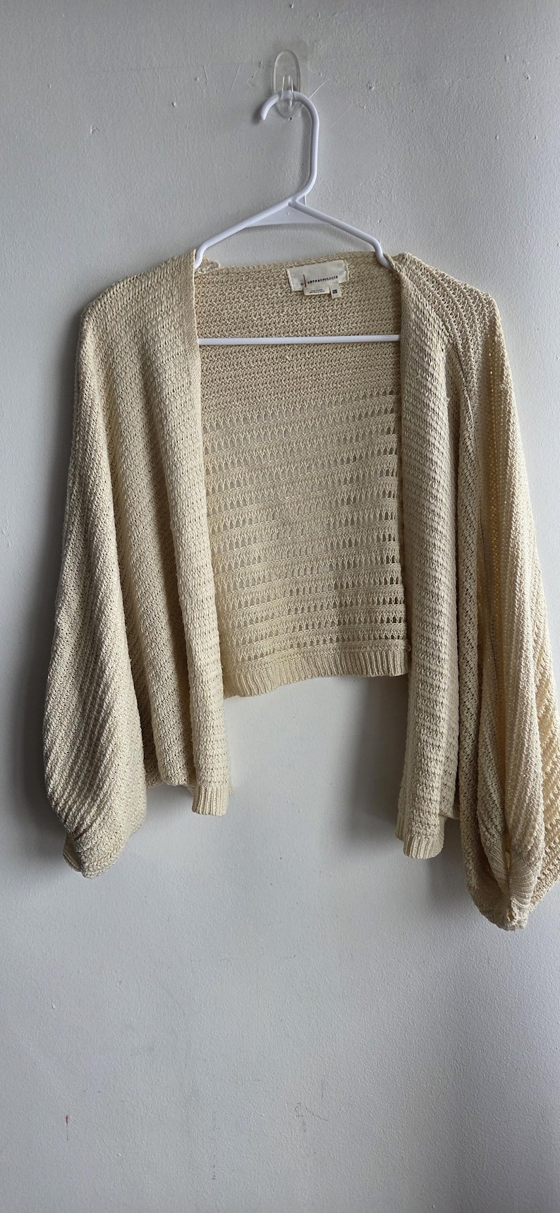 May include: A cream-colored, open-front cardigan with long, voluminous sleeves. The knitwear features a textured pattern and is displayed on a white hanger against a white wall. The cardigan is a lightweight, casual garment.