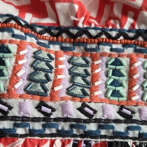 May include: Close-up of a vibrant textile with detailed embellishments. The fabric displays a red and white geometric print, with a beaded section in orange, blue, and purple hues. The design suggests a bohemian or ethnic style.