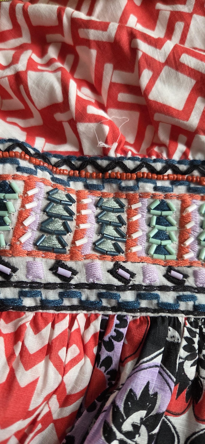 May include: Close-up of a vibrant textile with detailed embellishments. The fabric displays a red and white geometric print, with a beaded section in orange, blue, and purple hues. The design suggests a bohemian or ethnic style.