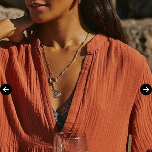 May include: A woman wearing an orange-red dress with a V-neck and a layered design. She is wearing a gold necklace and hoop earrings. She is holding a glass of water. The website editorialist.com is visible.