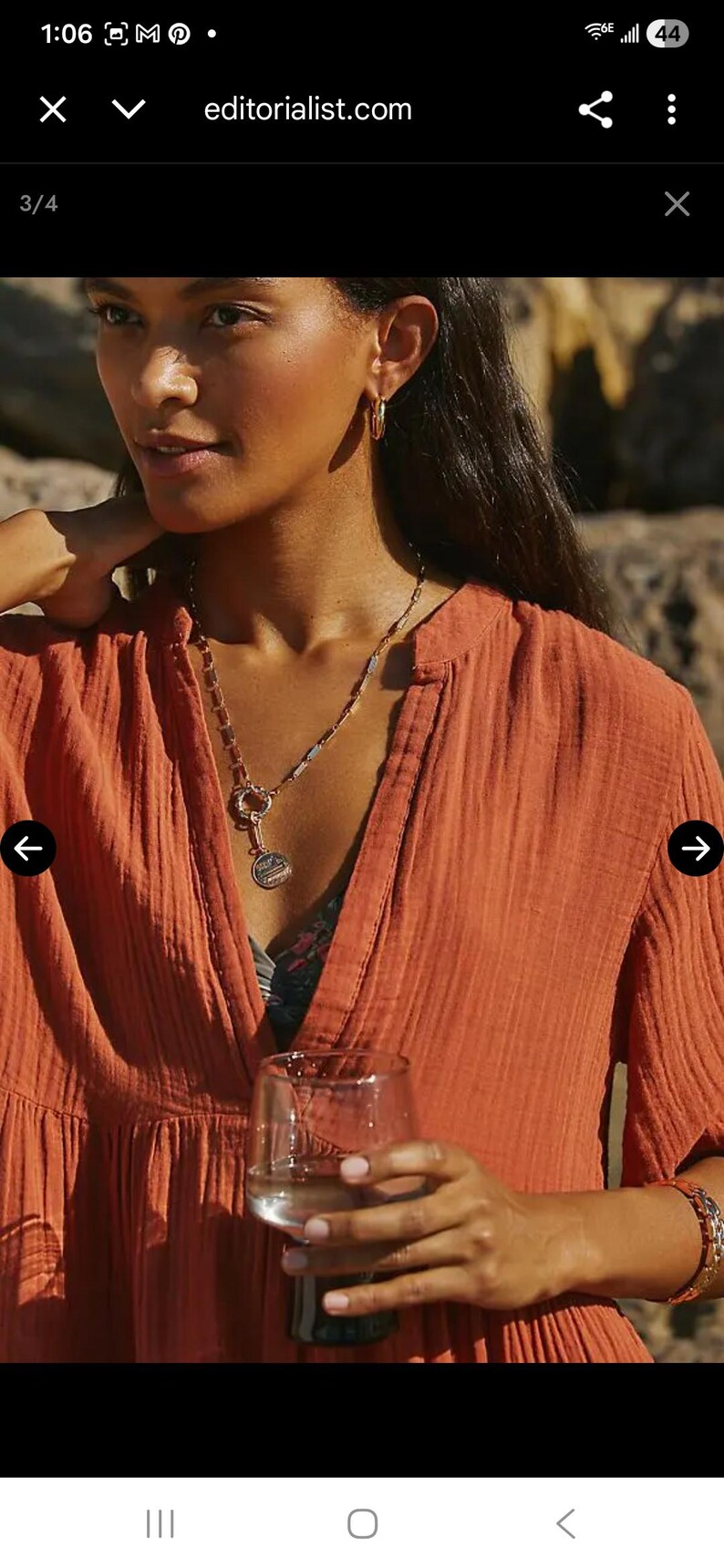 May include: A woman wearing an orange-red dress with a V-neck and a layered design. She is wearing a gold necklace and hoop earrings. She is holding a glass of water. The website editorialist.com is visible.