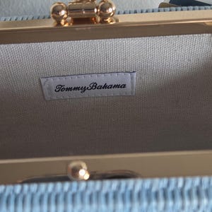 Puede incluir: Un bolso de mano tejido en color azul claro con un marco de metal dorado y un cierre decorativo en forma de concha. El interior est&aacute; forrado con una tela clara que incluye una etiqueta rectangular blanca con el texto negro Tommy Bahama.