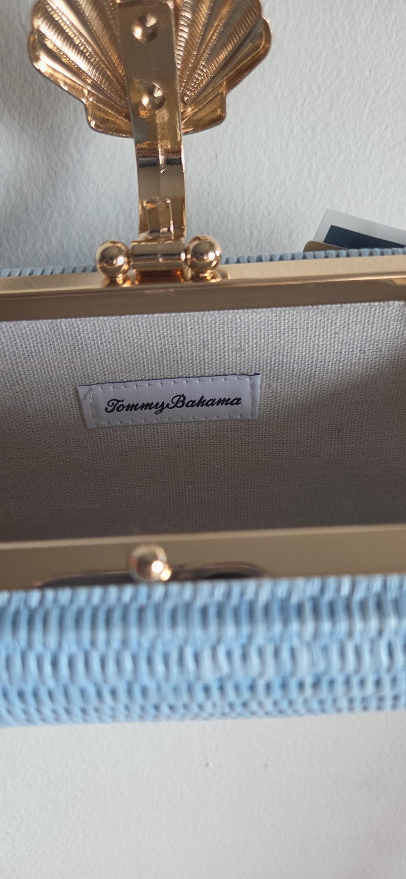 Puede incluir: Un bolso de mano tejido en color azul claro con un marco de metal dorado y un cierre decorativo en forma de concha. El interior est&aacute; forrado con una tela clara que incluye una etiqueta rectangular blanca con el texto negro Tommy Bahama.
