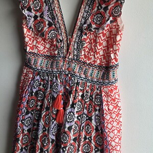 May include: A patterned romper with a V-neck and frilled sleeves. The fabric displays a floral design in red, black, and purple, with geometric details. A red tassel hangs from the waist.