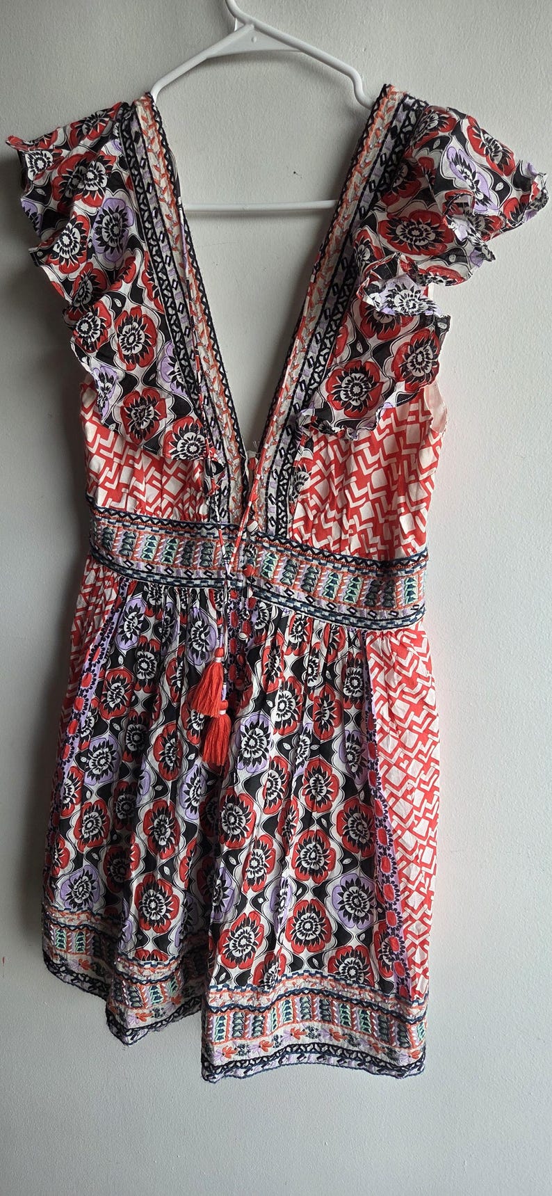 May include: A patterned romper with a V-neck and frilled sleeves. The fabric displays a floral design in red, black, and purple, with geometric details. A red tassel hangs from the waist.