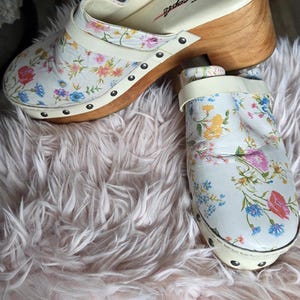 May include: White clogs with a floral pattern featuring pink, blue, and yellow flowers. The clogs have a wooden heel and the brand name "free people" is visible on the insole.