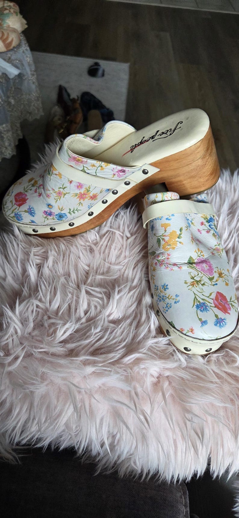 May include: White clogs with a floral pattern featuring pink, blue, and yellow flowers. The clogs have a wooden heel and the brand name "free people" is visible on the insole.