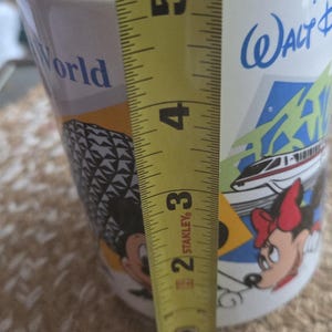 May include: White ceramic mug with a colorful design featuring Mickey and Minnie Mouse, a monorail, and the words "Walt Disney World." The mug is approximately 4 inches tall, as measured by a yellow tape measure.