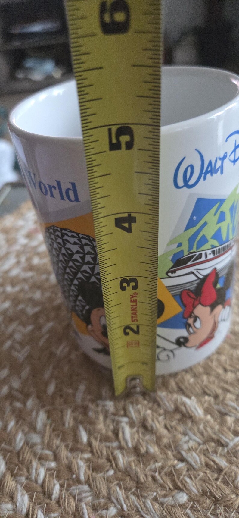 May include: White ceramic mug with a colorful design featuring Mickey and Minnie Mouse, a monorail, and the words "Walt Disney World." The mug is approximately 4 inches tall, as measured by a yellow tape measure.