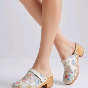 May include: Floral-print clogs with wooden heels. The clogs feature a white base adorned with a colorful floral pattern, a cream-colored strap, and a wooden heel. The soles are trimmed with silver studs. These shoes are a stylish addition to any wardrobe.