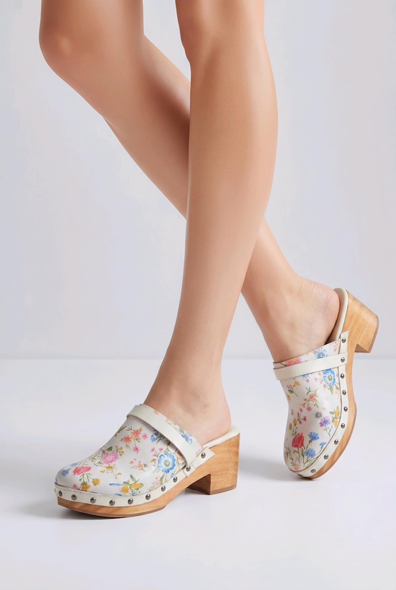 May include: Floral-print clogs with wooden heels. The clogs feature a white base adorned with a colorful floral pattern, a cream-colored strap, and a wooden heel. The soles are trimmed with silver studs. These shoes are a stylish addition to any wardrobe.