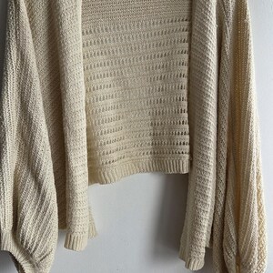 May include: Cream-colored open-front cardigan with long, voluminous sleeves. The knitwear features a textured pattern and is displayed on a white hanger against a white wall. The cardigan has a cropped length.